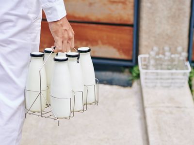 A milkman's arm holding a 6-pack of milk bottles as he walks towards a front door.